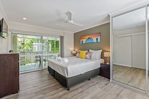 Un dormitorio con cama, mesitas de noche, ventilador de techo, un cuadro y un balcón con vistas al verdor.