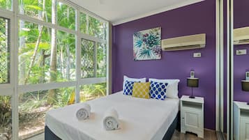 A bedroom with a bed, two nightstands, a wall-mounted air conditioner, and a window with a view of greenery.