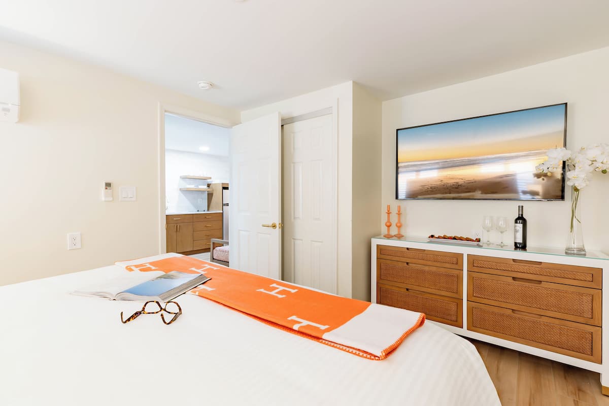 A hotel room with a bed, a bedside table with a book and glasses, a dresser, a painting on the wall, and a vase with flowers.