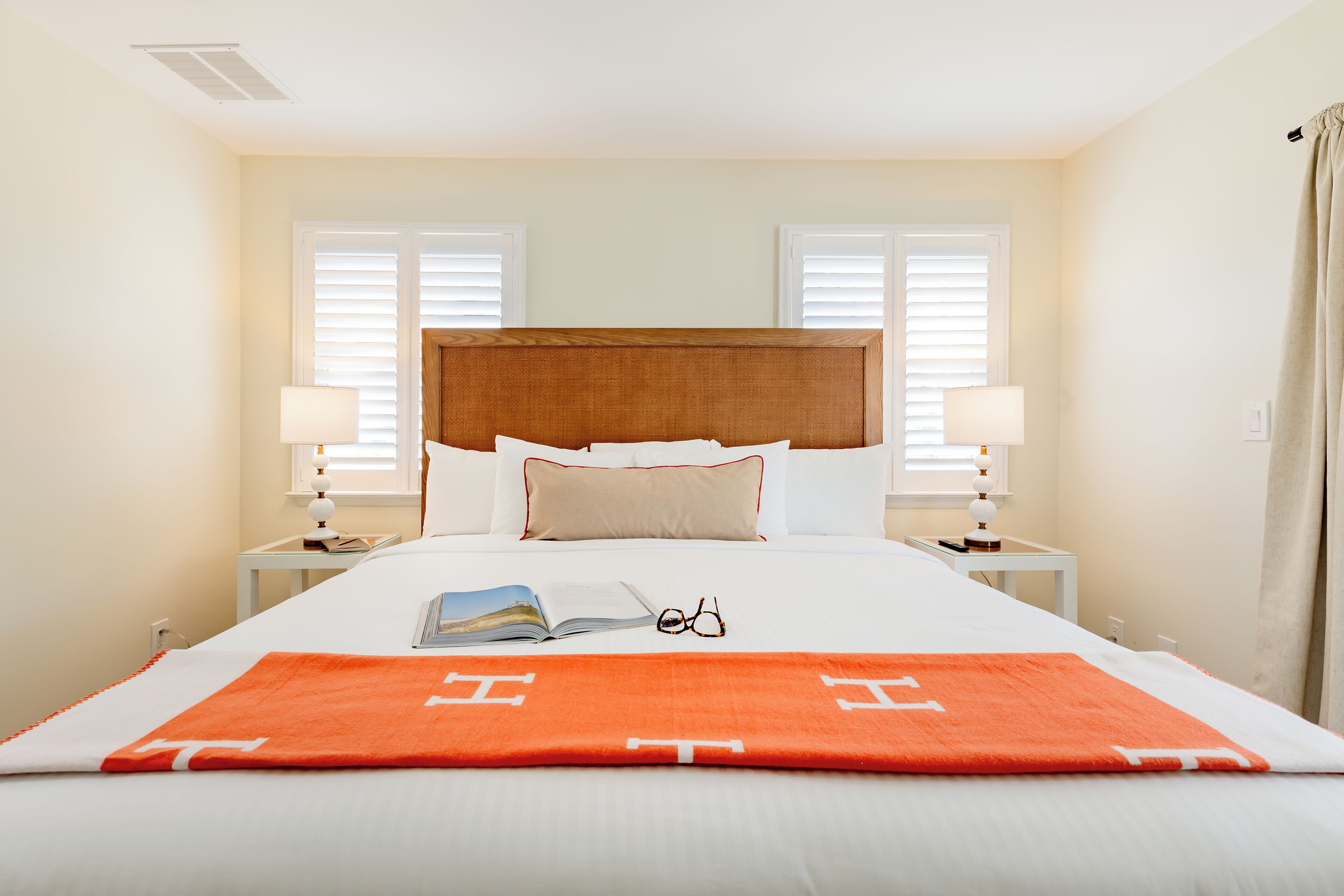 A neatly made bed with a white and orange blanket, two bedside lamps, and a book and glasses on the bed.
