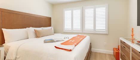 Una cama bien hecha con sábanas blancas y una manta naranja, una mesita de noche con un libro y gafas, y un aparador con un espejo.