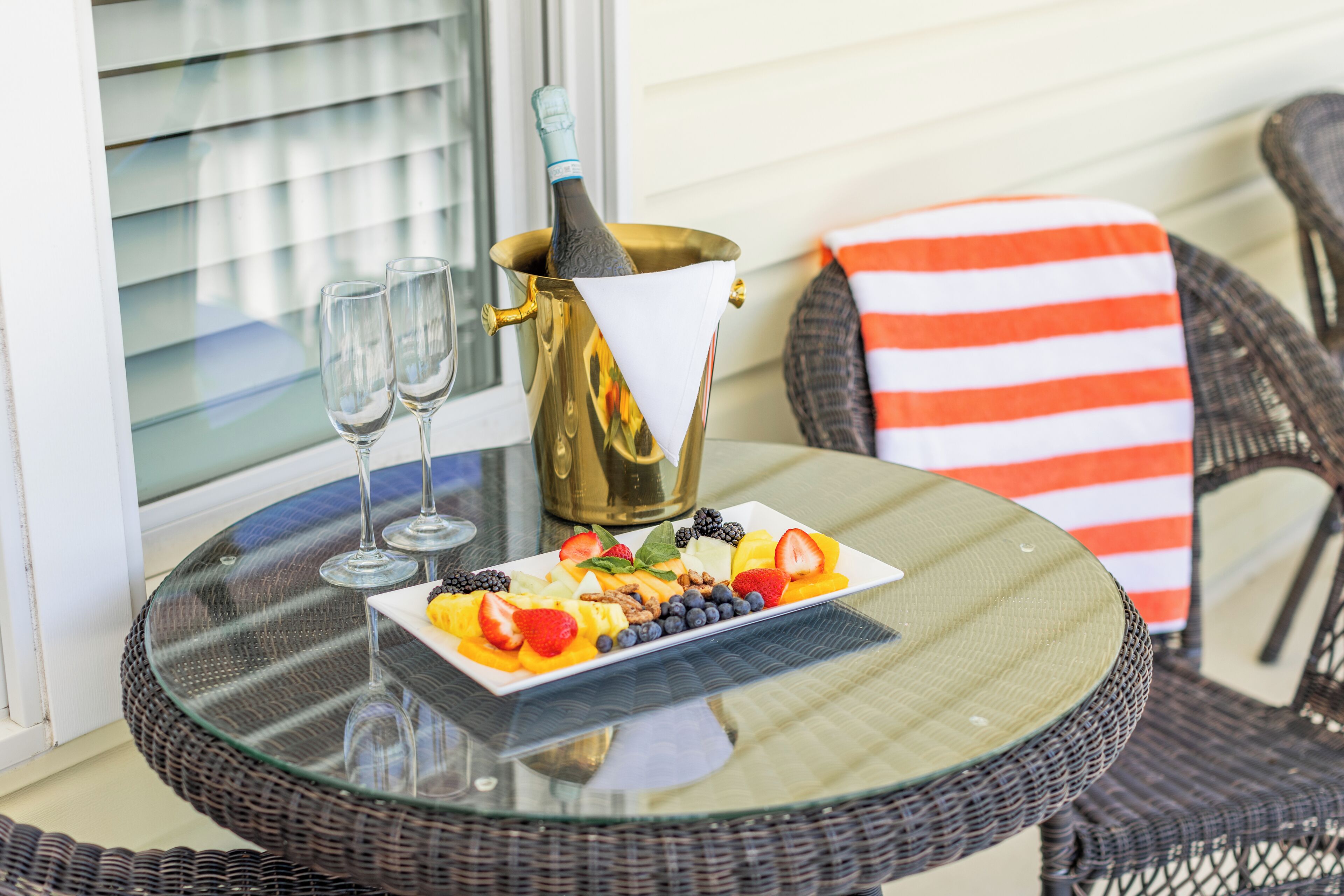 A glass table with a bottle of champagne, two glasses, and a plate of fruit.