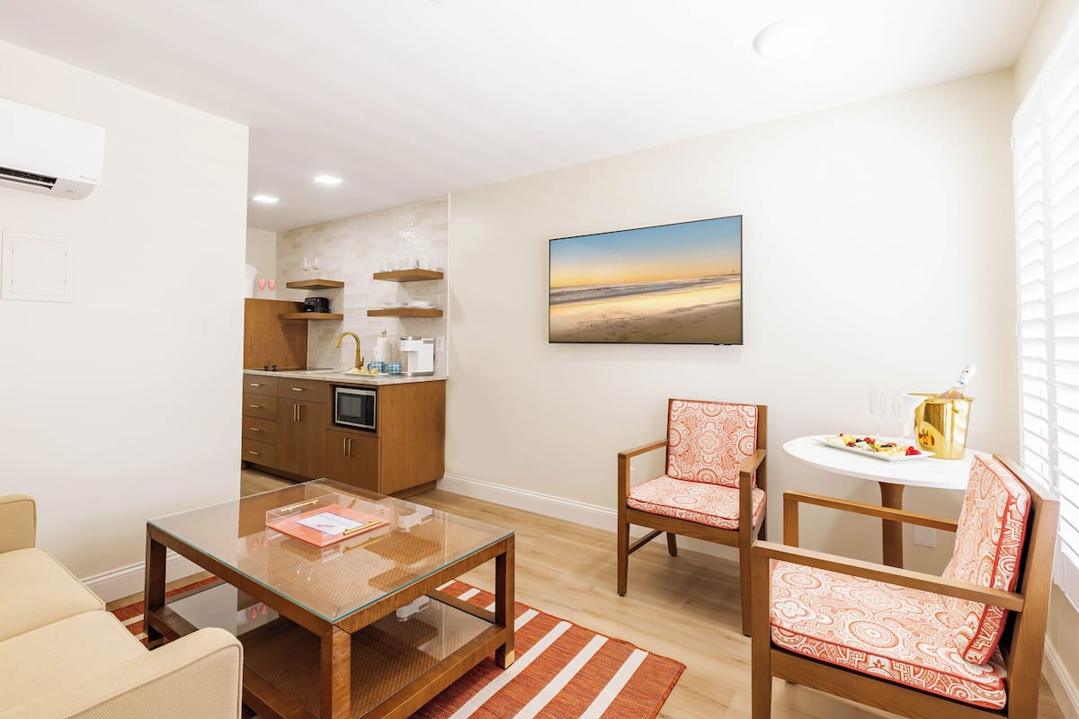 A modern living room with a glass coffee table, two patterned chairs, a sofa, and a kitchen area with wooden cabinets and a built-in oven.