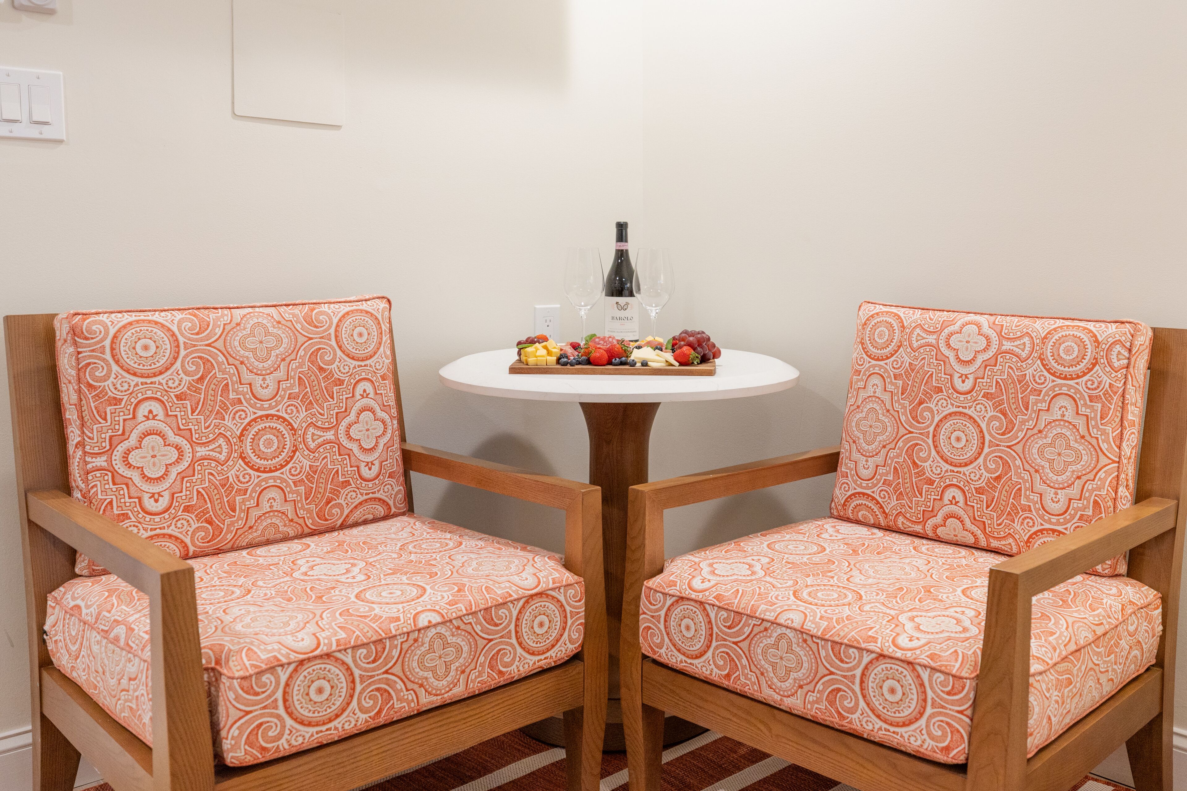 Una habitación con dos sillas de madera con cojines de estampado naranja, una mesita redonda con una botella de vino y copas, y una bandeja de fruta.