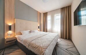 Habitación de hotel con una cama grande, cabecera, mesita de noche con lámpara, televisor montado en la pared y ventana con cortinas.