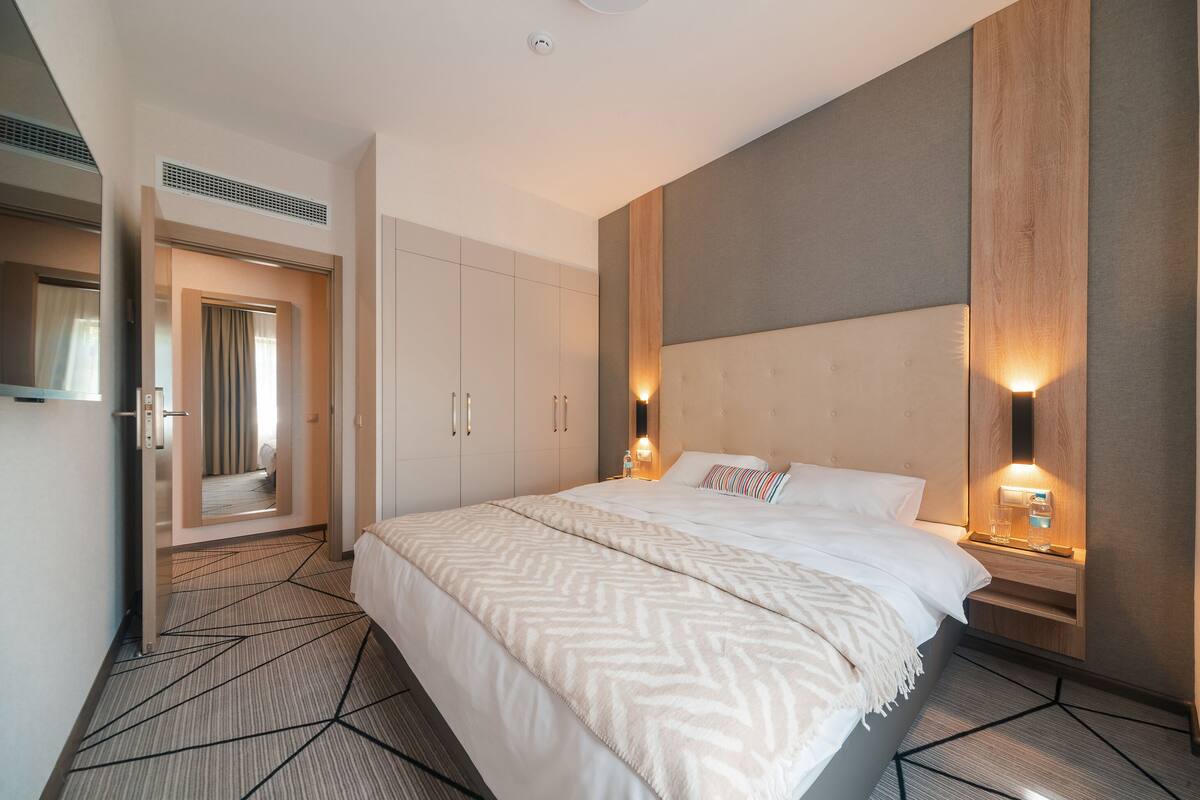 Una habitación de hotel moderna con una cama grande, un armario empotrado, una mesita de noche con lámpara y un espejo.