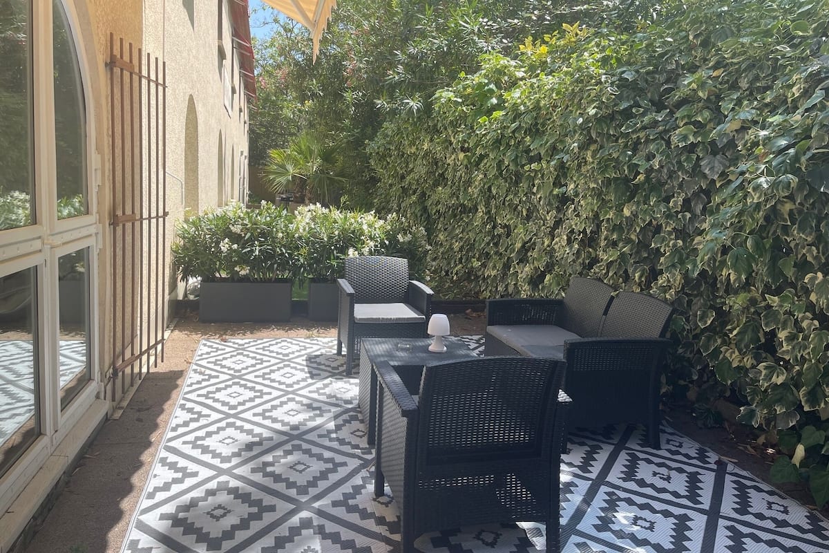 Un espace extérieur aménagé en terrasse, avec un sol à motifs, des meubles en osier noir et une grande haie touffue.