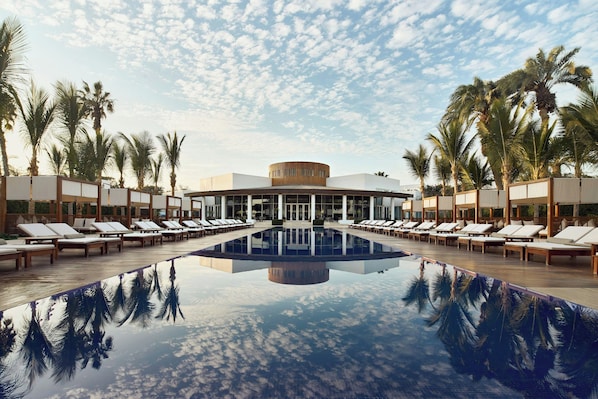 A pool with lounge chairs, palm trees, and a building with a circular central section.