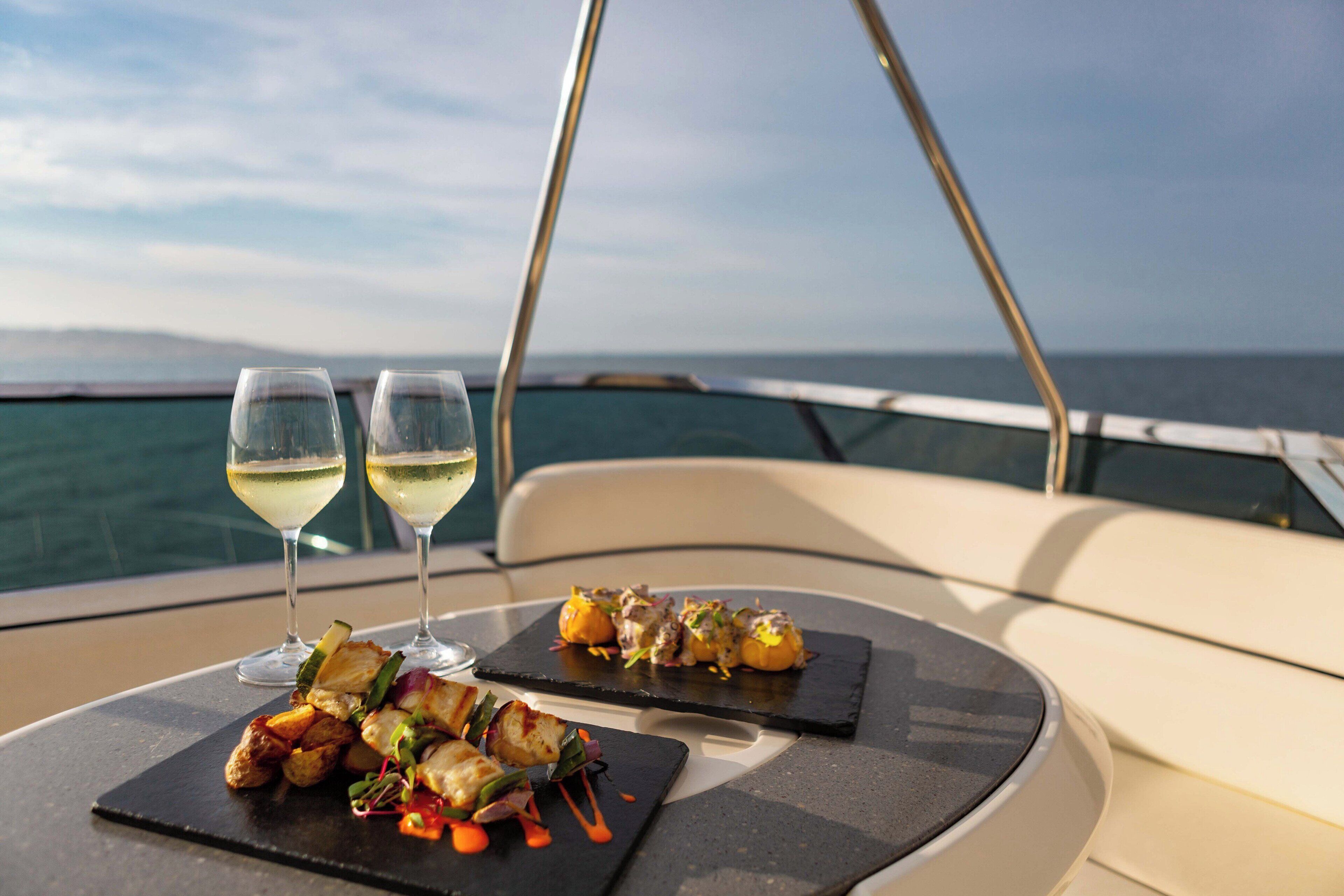 A table with two glasses of white wine and a plate of food on a boat.