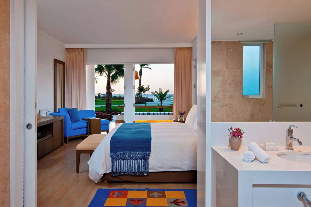 Une chambre d’hôtel avec un lit, un canapé bleu, une petite table, une salle de bain avec un lavabo et un miroir, et une vue sur un paysage verdoyant.