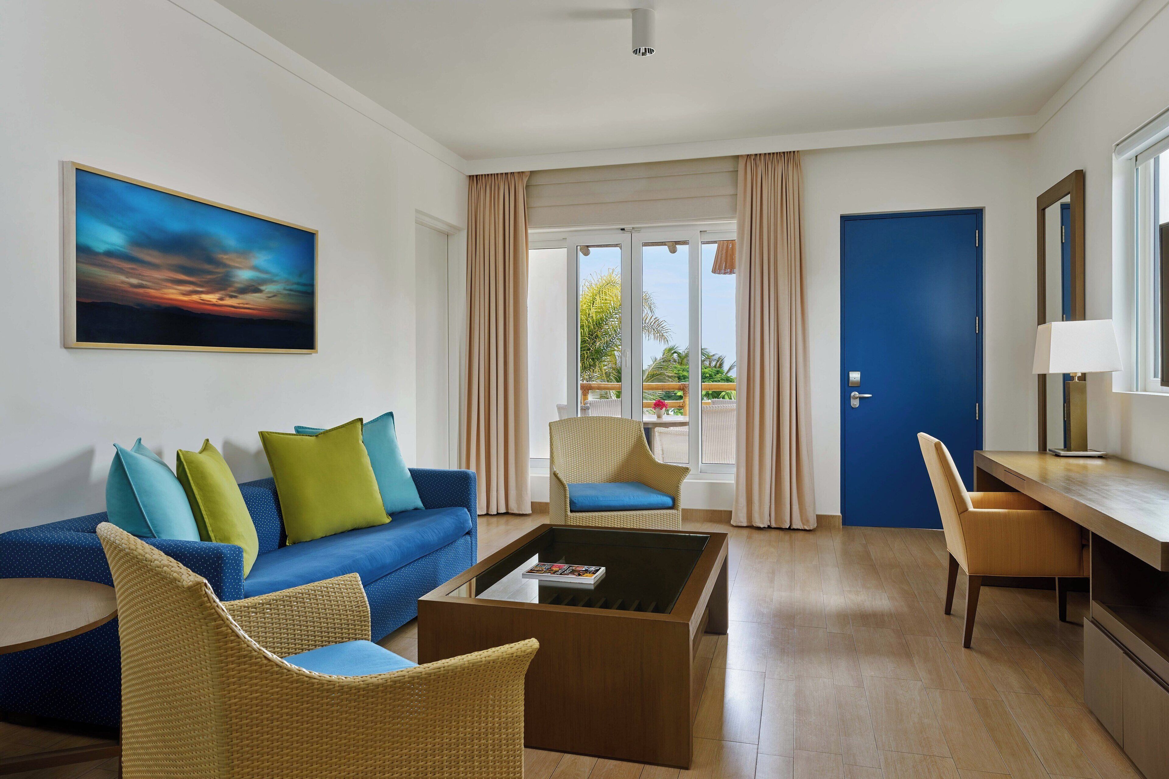 Een moderne woonkamer met een blauwe bank, rieten stoelen, een salontafel en een groot raam met gordijnen.