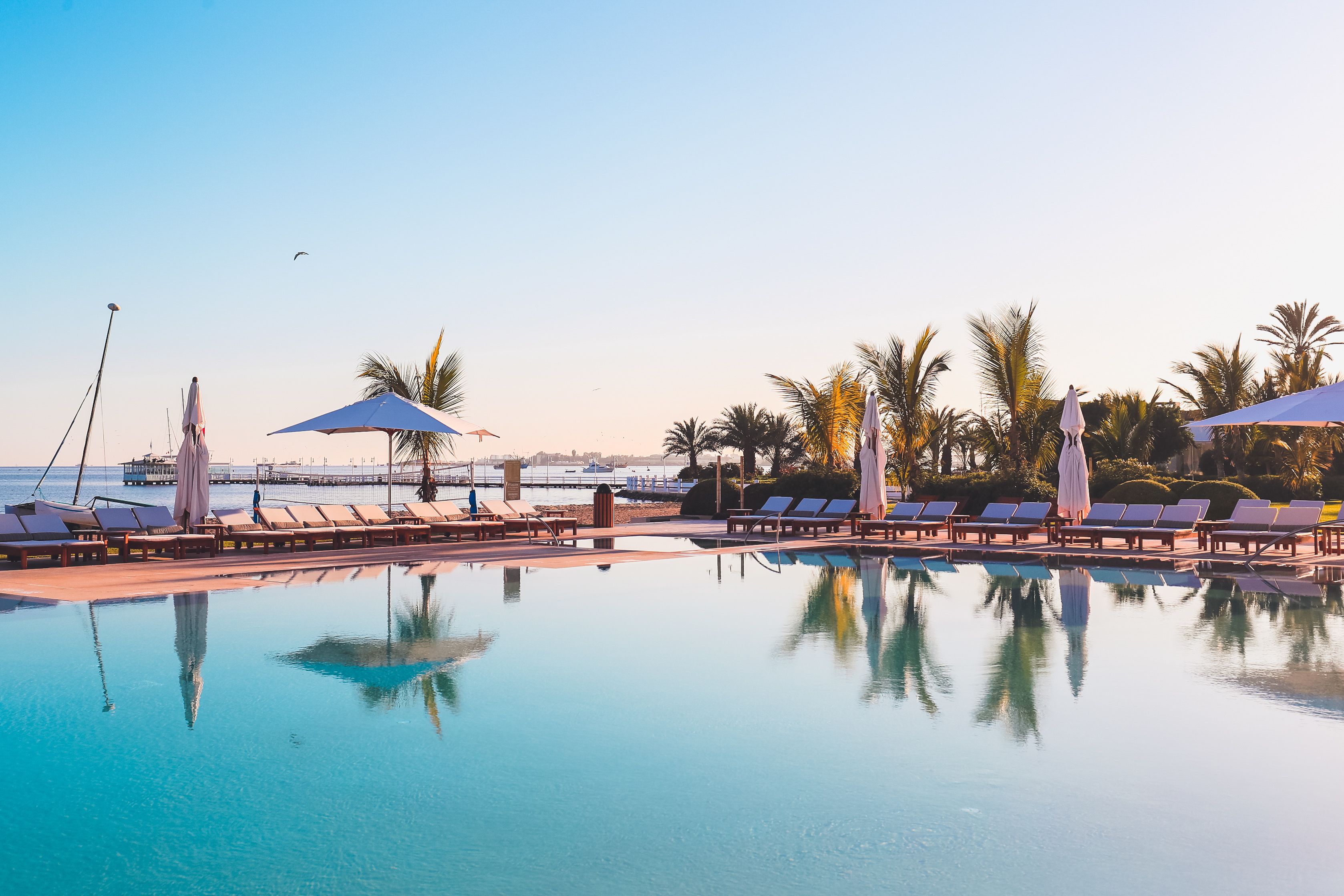 Zona de piscina con sillones de playa y sombrillas, cielo despejado y vista al mar.