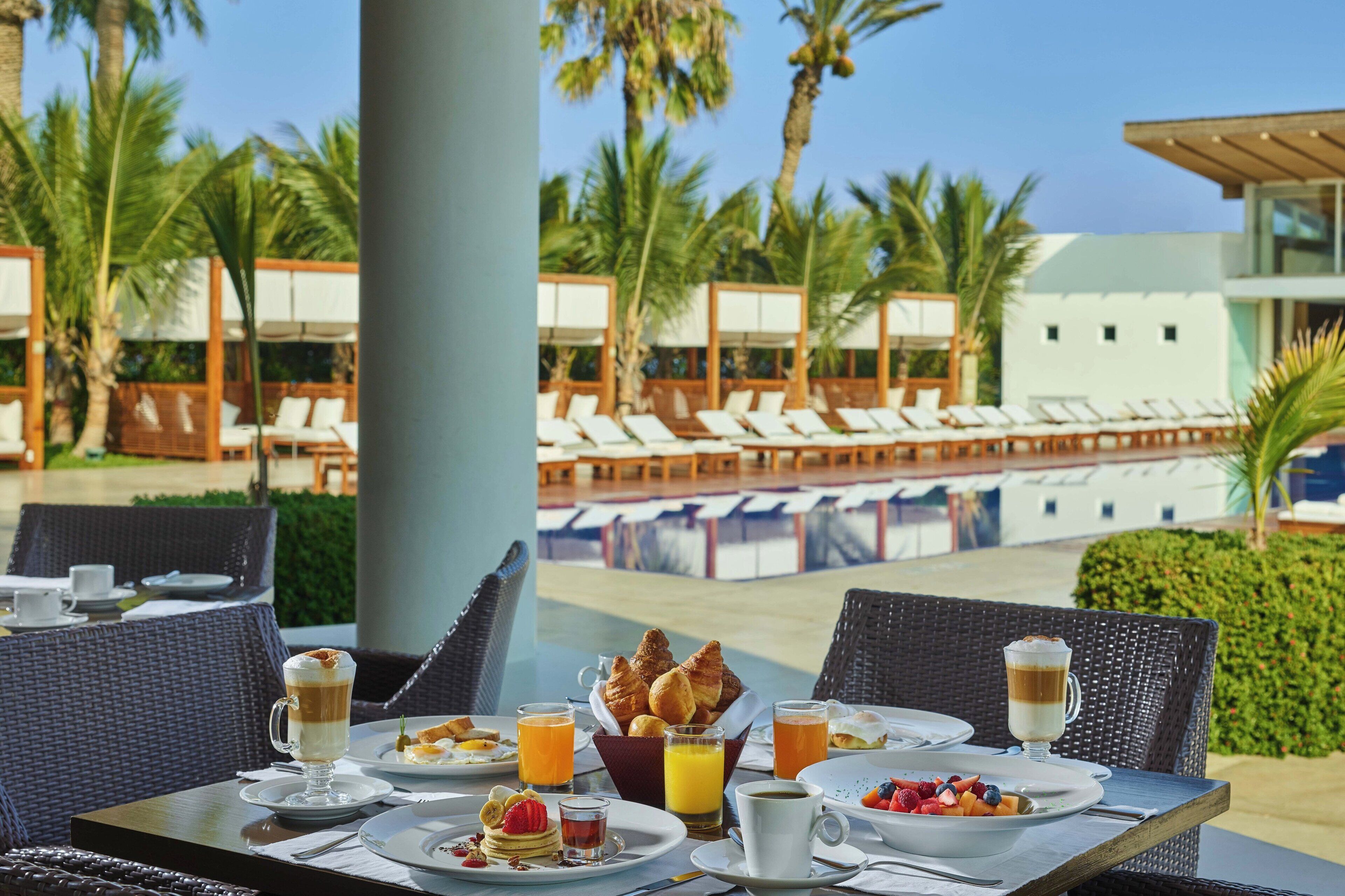 A breakfast table with various food items and drinks, overlooking a pool area with lounge chairs and palm trees.