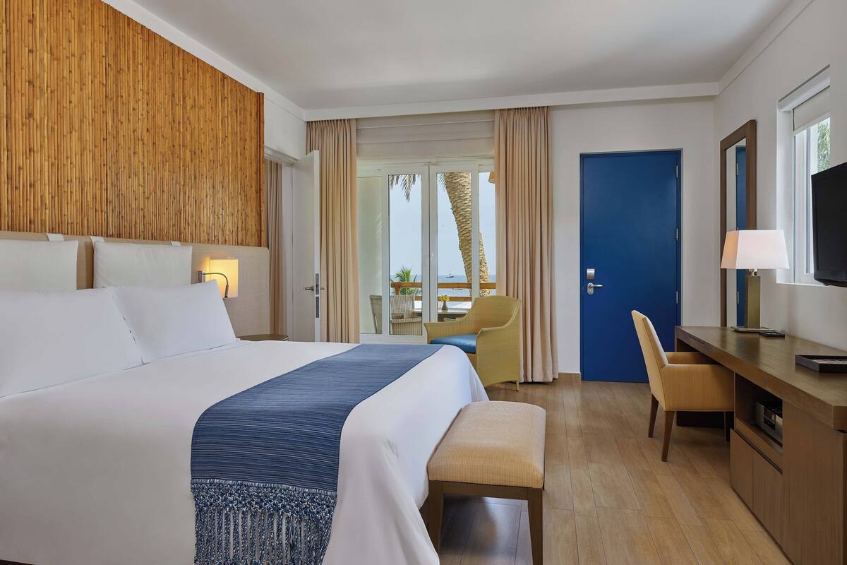 Une chambre d’hôtel avec un grand lit, un bureau avec une chaise, une télévision et une porte donnant sur un balcon avec vue sur la mer.