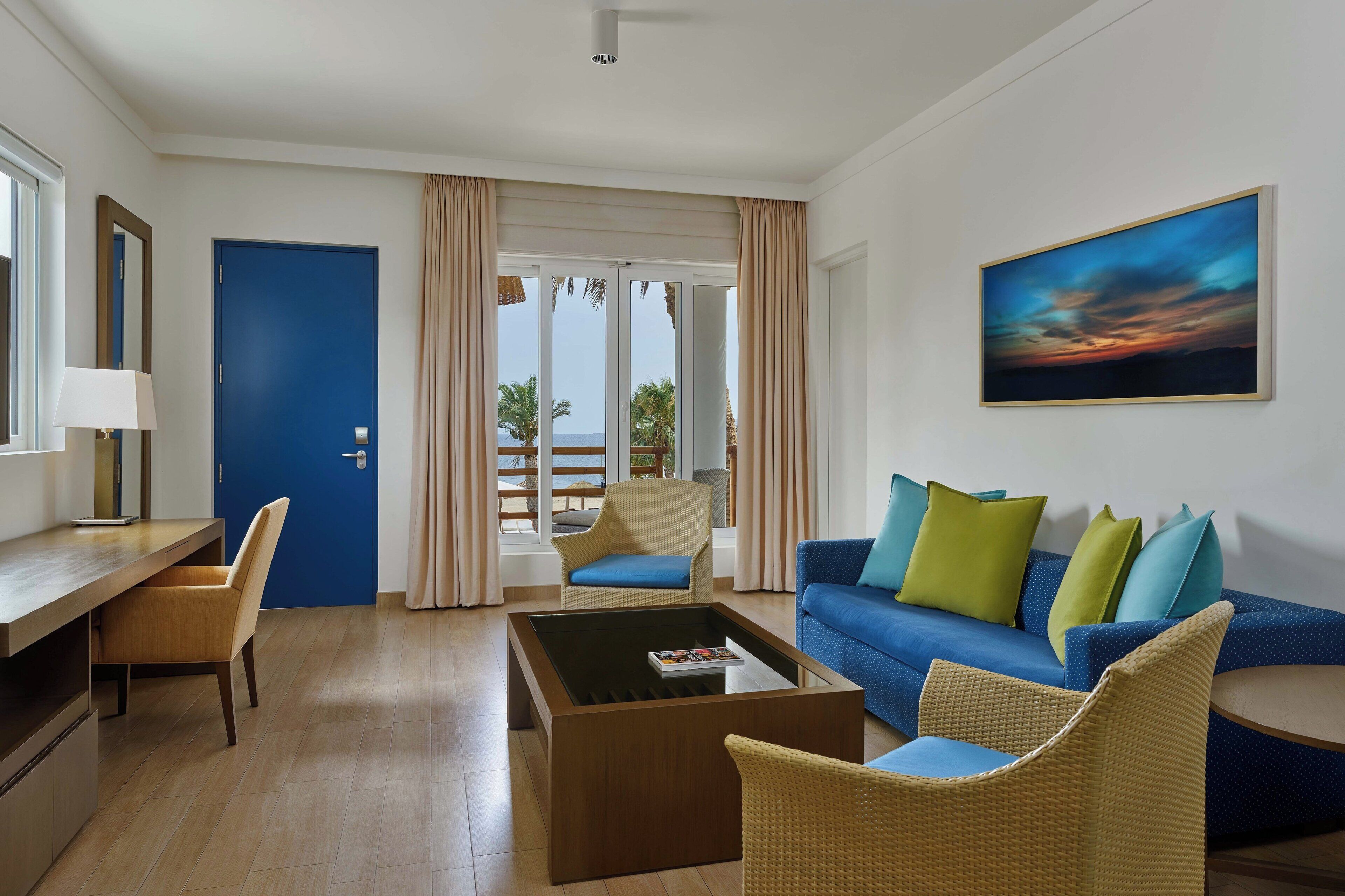 A hotel room with a blue door, a sofa with multiple cushions, a coffee table, a chair, and a painting on the wall.
