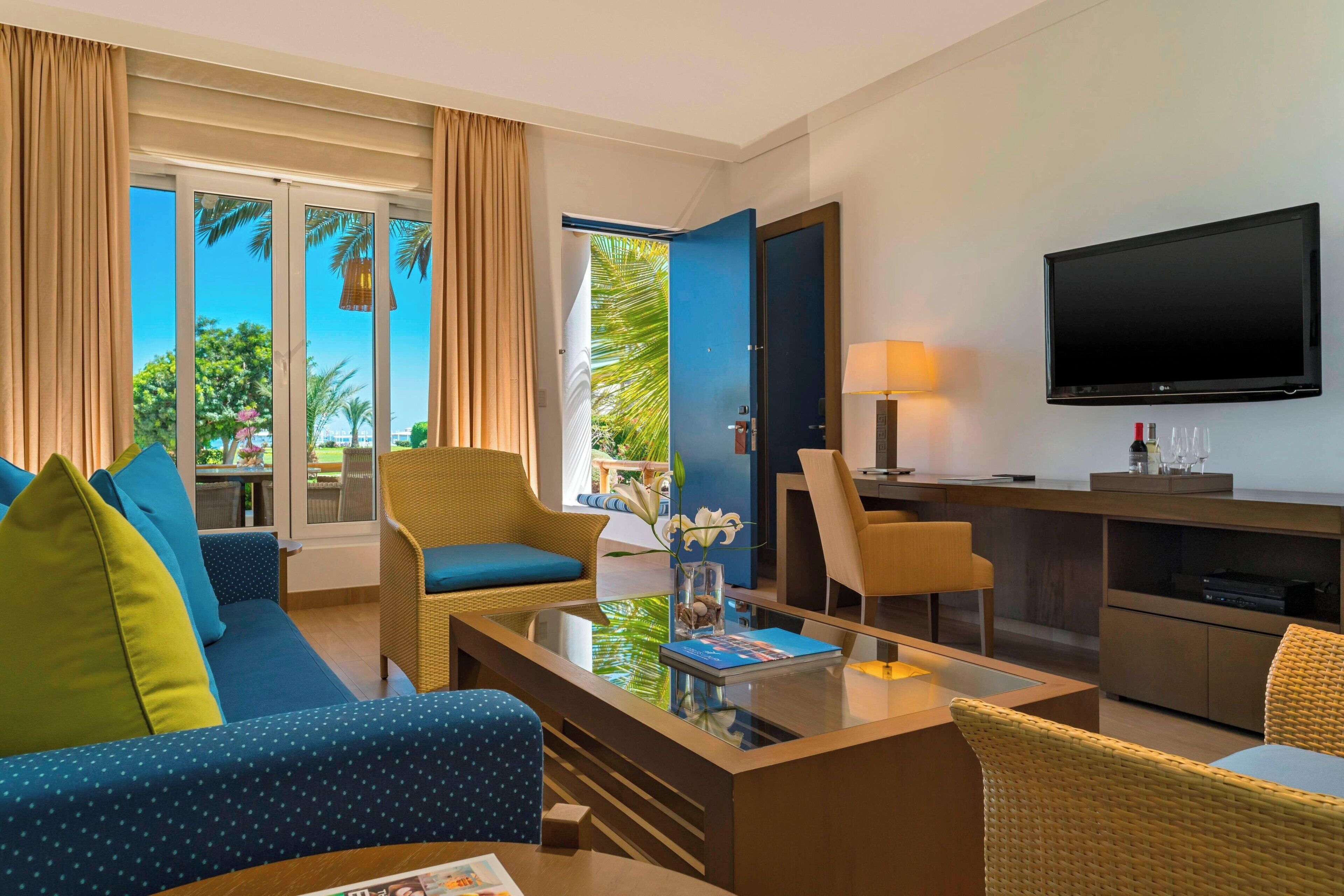 A modern hotel room with a large TV, comfortable seating, a glass coffee table, and a view of palm trees through the sliding glass door.