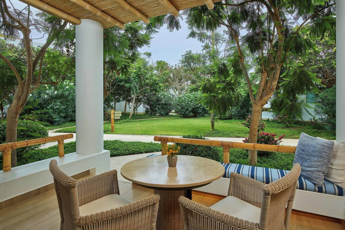 Une terrasse couverte, aménagée avec des meubles en osier, une table ronde et offrant une vue sur un jardin bien entretenu.
