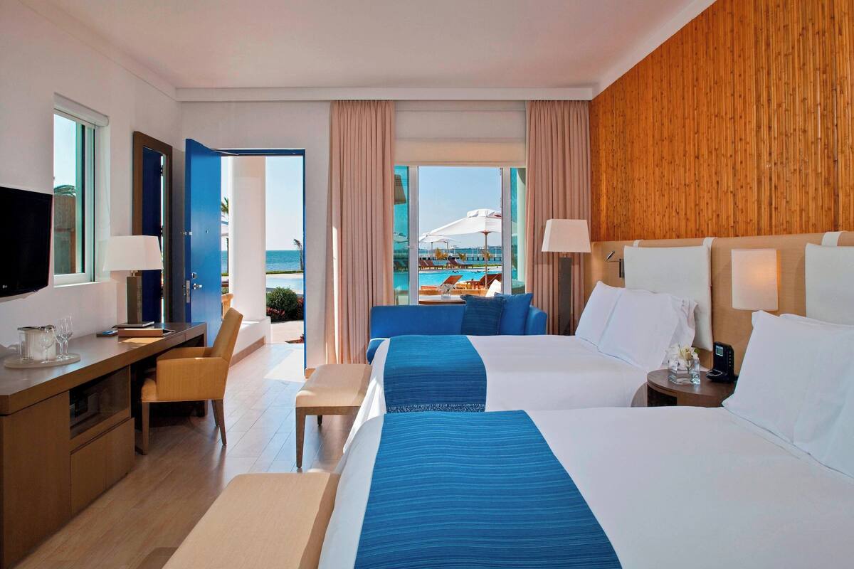 Une chambre d’hôtel avec deux lits, un bureau, une chaise, une télévision et une vue sur la piscine et l’océan.