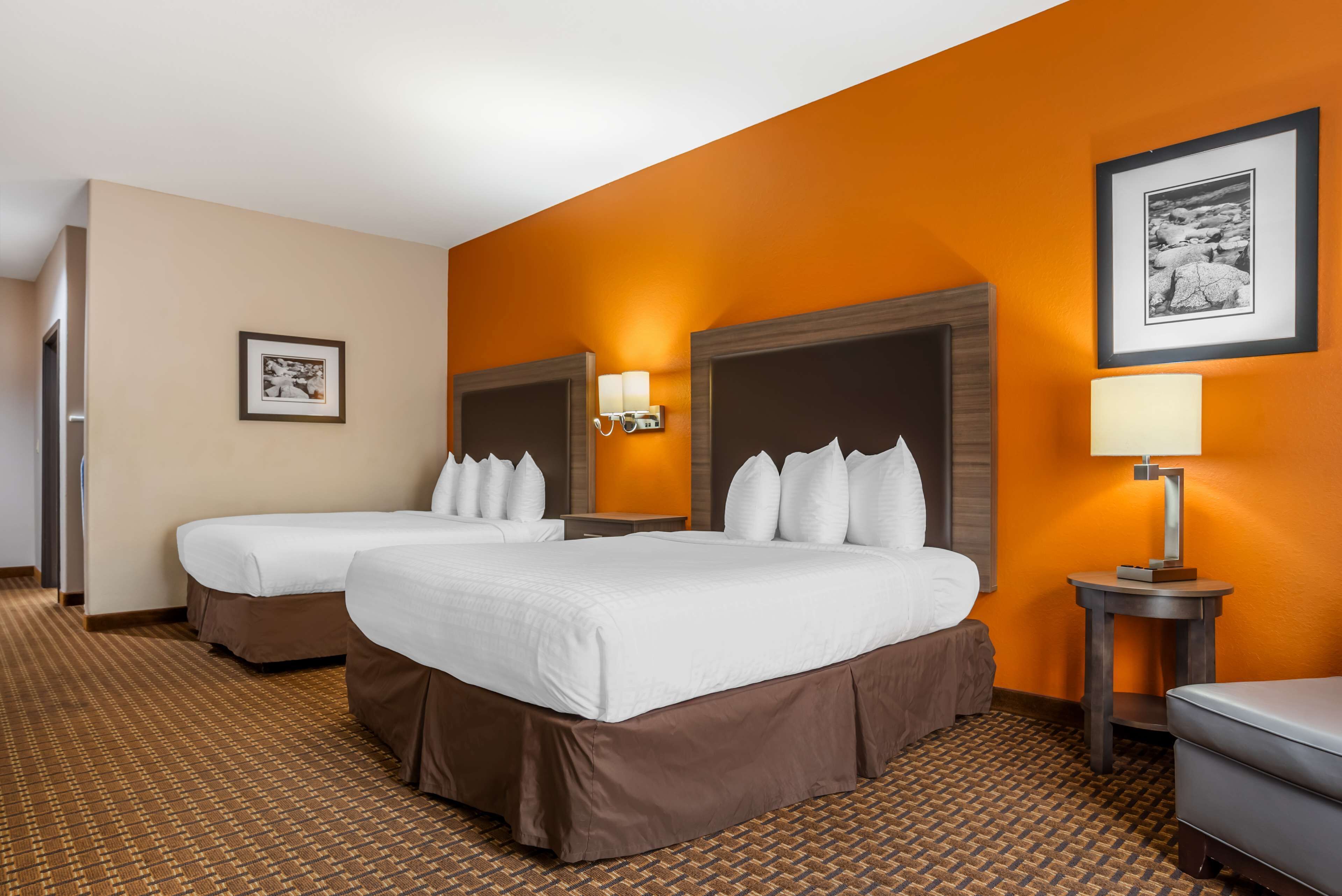A hotel room with two beds, a brown carpet, a wall with an orange accent, and a framed picture on the wall.
