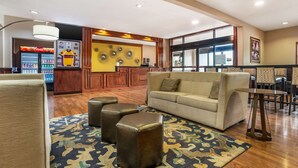 Un hall de hotel con un sofá, otomanas, un tapete estampado, una recepción, un refrigerador y una máquina expendedora.