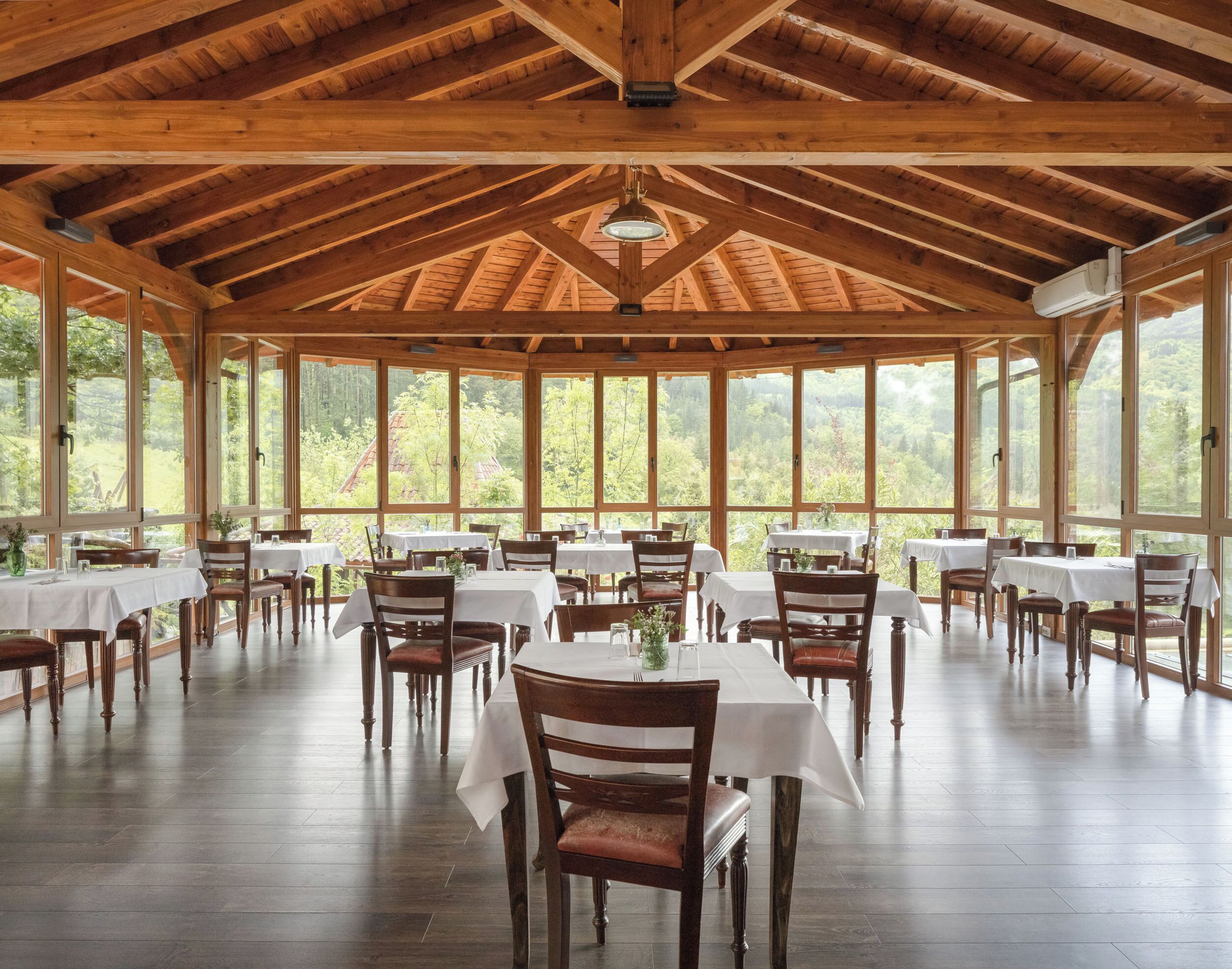 A spacious dining area with wooden beams and large windows offering a view of the forest. Tables are set with white tablecloths and chairs.