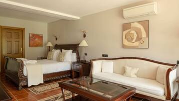 A hotel room with a bed, a sofa, a coffee table, a nightstand, a lamp, and an air conditioning unit.