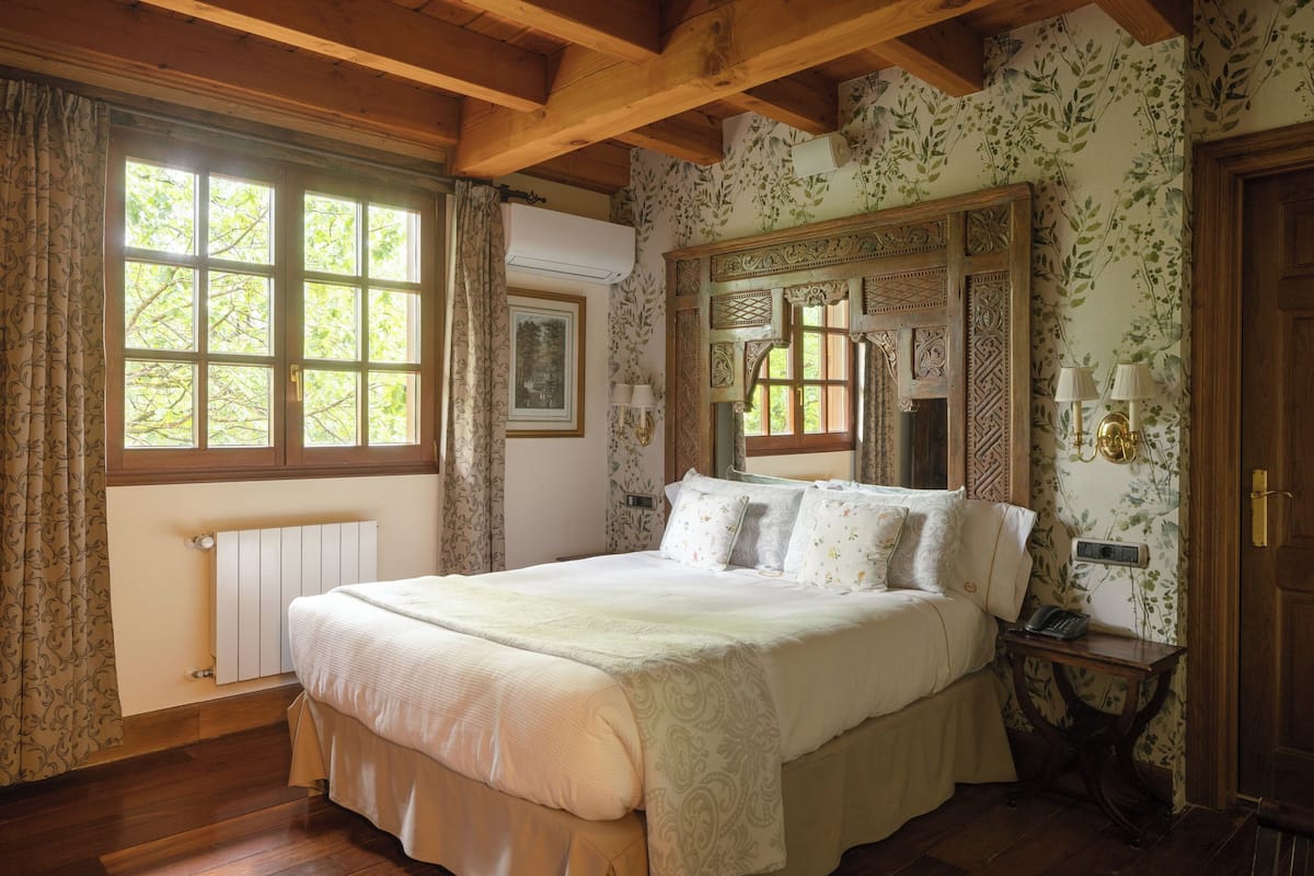 A bedroom with a large bed, wooden beams, a radiator, a window with curtains, a mirror, and a bedside table with a lamp.