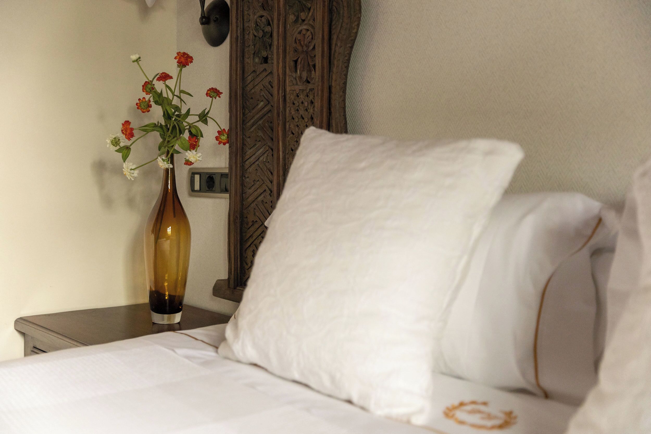 A neatly made bed with white linens, a white pillow, and a vase with flowers on a bedside table.