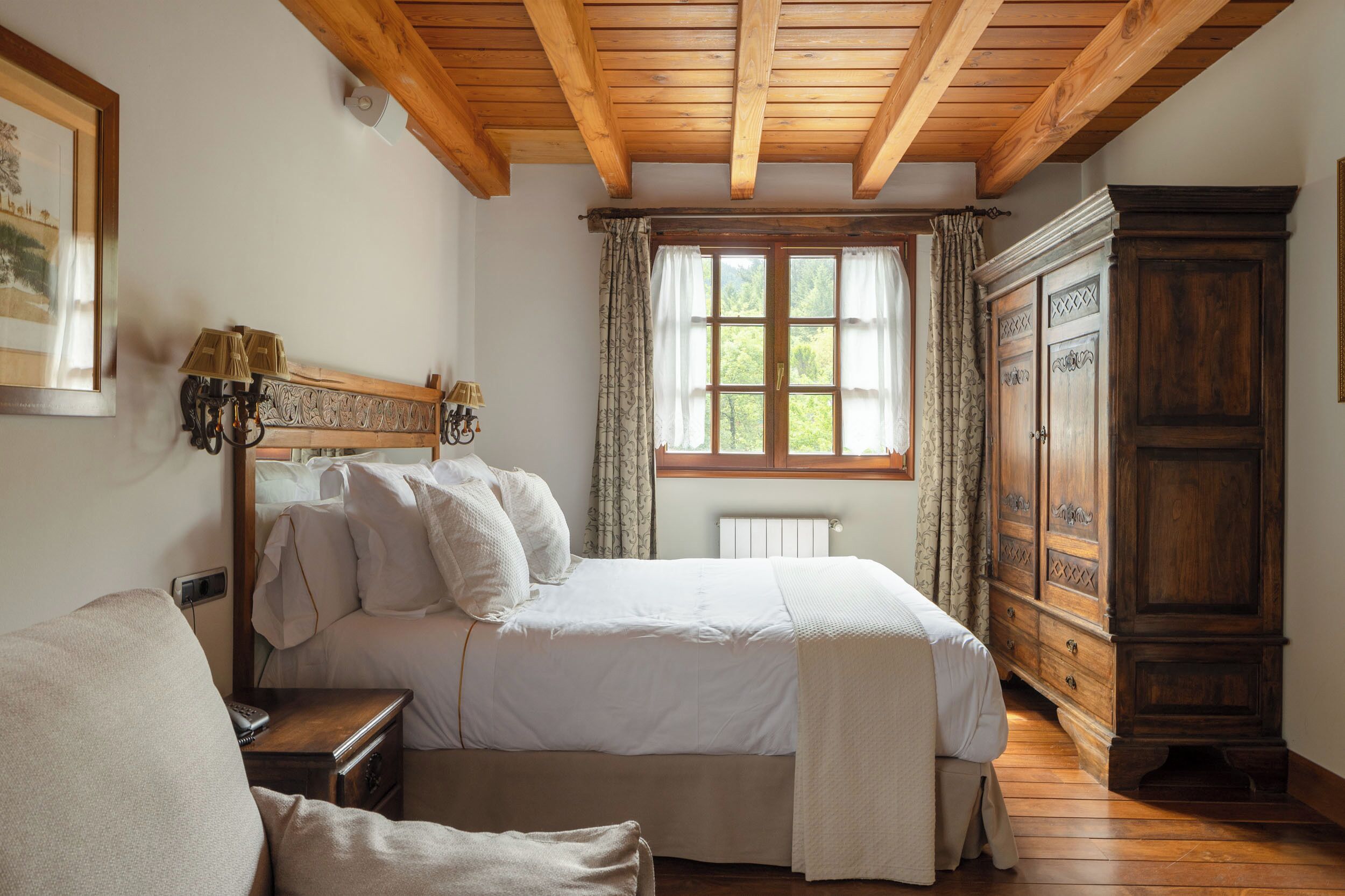 Un dormitorio con techo de madera, una cama grande con ropa blanca, una mesita de noche con lámpara, un armario de madera y una ventana con cortinas.