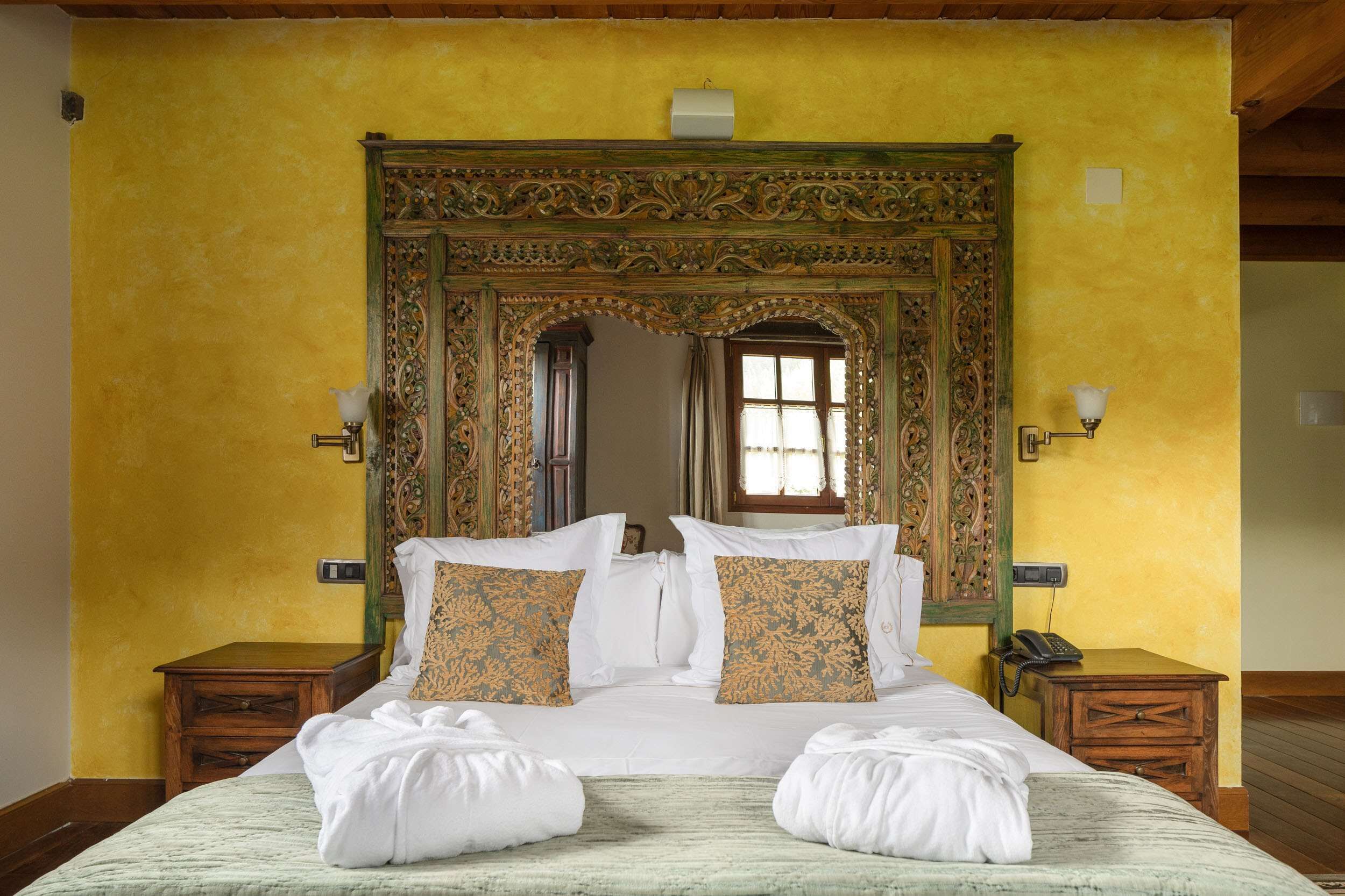 A hotel room with a large bed, ornate wooden headboard, two bedside tables, and a window with curtains.