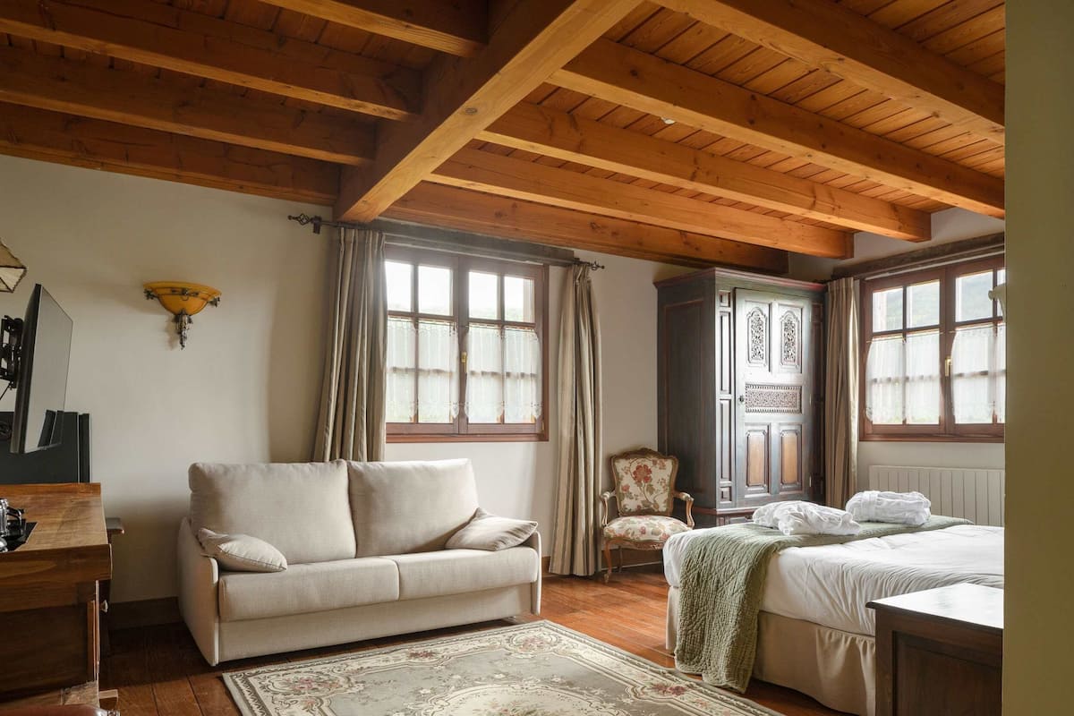 A room with a wooden ceiling, a sofa, a chair, a bed, and a window with curtains.