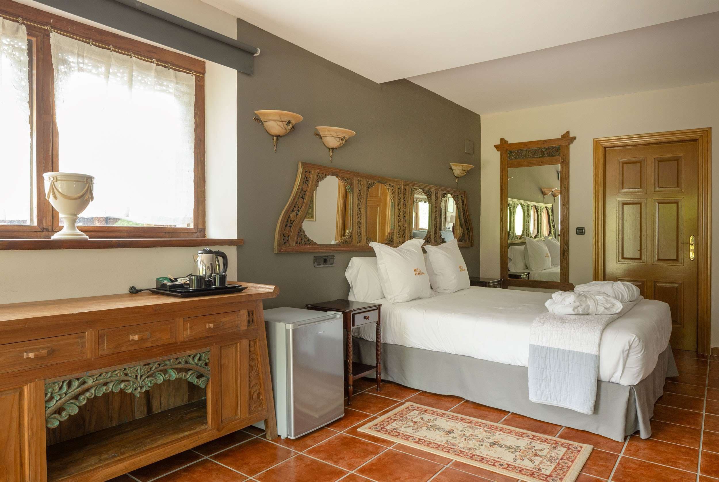 A bedroom with a bed, a dresser, a small refrigerator, and a window with a curtain.
