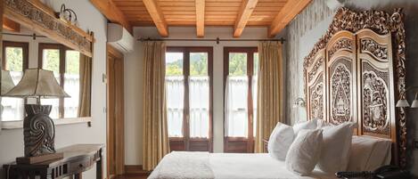 Una habitación de hotel con techo de madera, una cama grande con sábanas blancas, una mesita de noche de madera, un escritorio con lámpara y un espejo grande y decorativo.