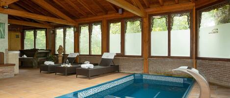 Una piscina interior con borde de azulejos, vigas de madera en el techo y una zona de estar con chimenea.