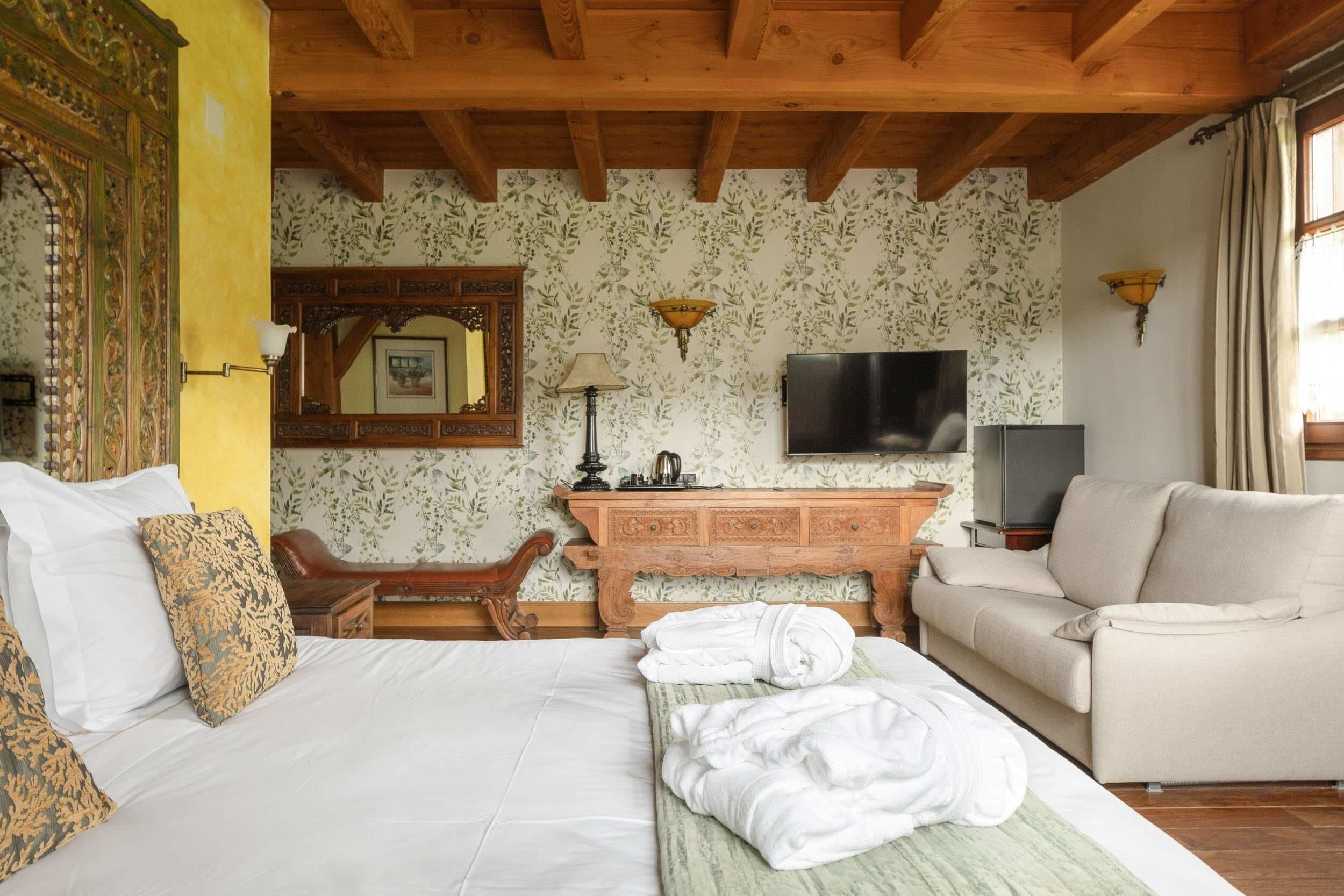 Habitación de hotel con techo de madera, cama con ropa blanca, mesita de noche de madera, sofá beige, televisor de pantalla plana y papel tapiz floral.