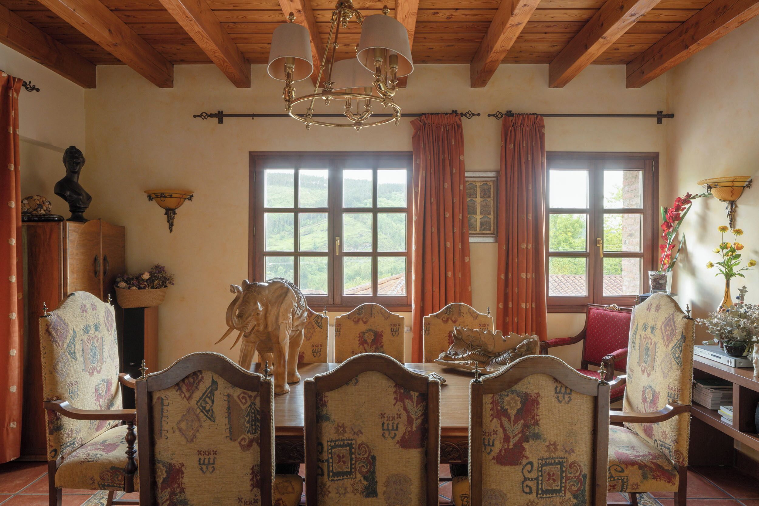 A dining area with a wooden ceiling, patterned chairs, a chandelier, and large windows with curtains.