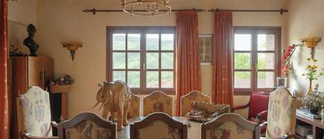 A dining area with a wooden ceiling, patterned chairs, a chandelier, and large windows with curtains.