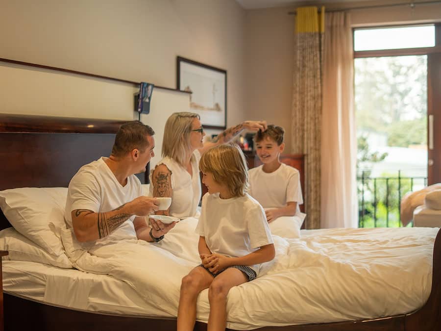 A family of four on a bed with a wooden headboard, a window with curtains, and a bedside table with a lamp.