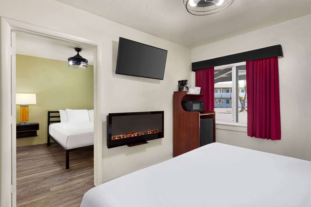 A hotel room with a bed, a flat-screen TV mounted on the wall, a desk with a lamp, a microwave, and a window with red curtains.