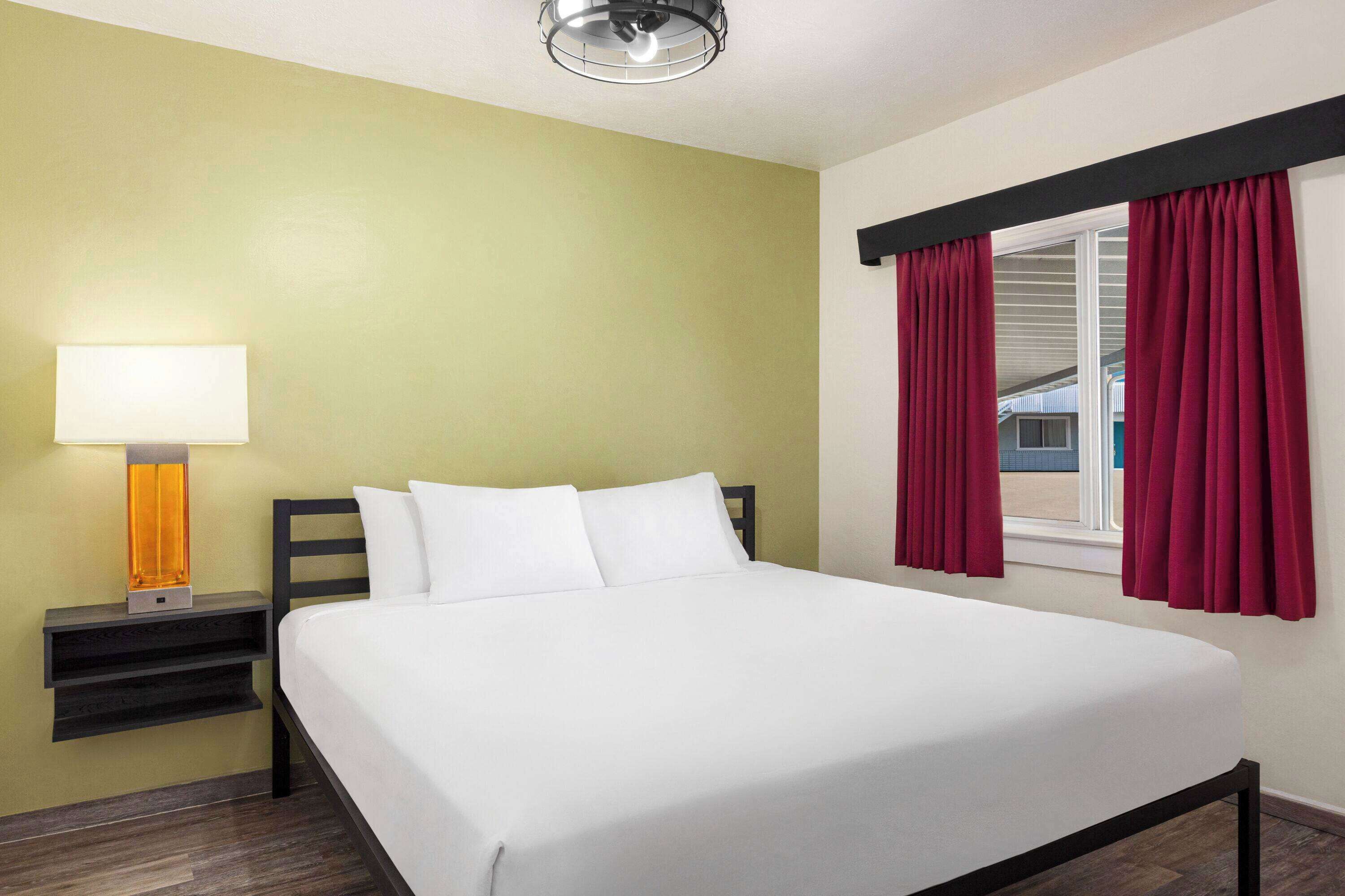 A neatly made bed in a room with a green wall, a bedside table with a lamp, and red curtains on a window.