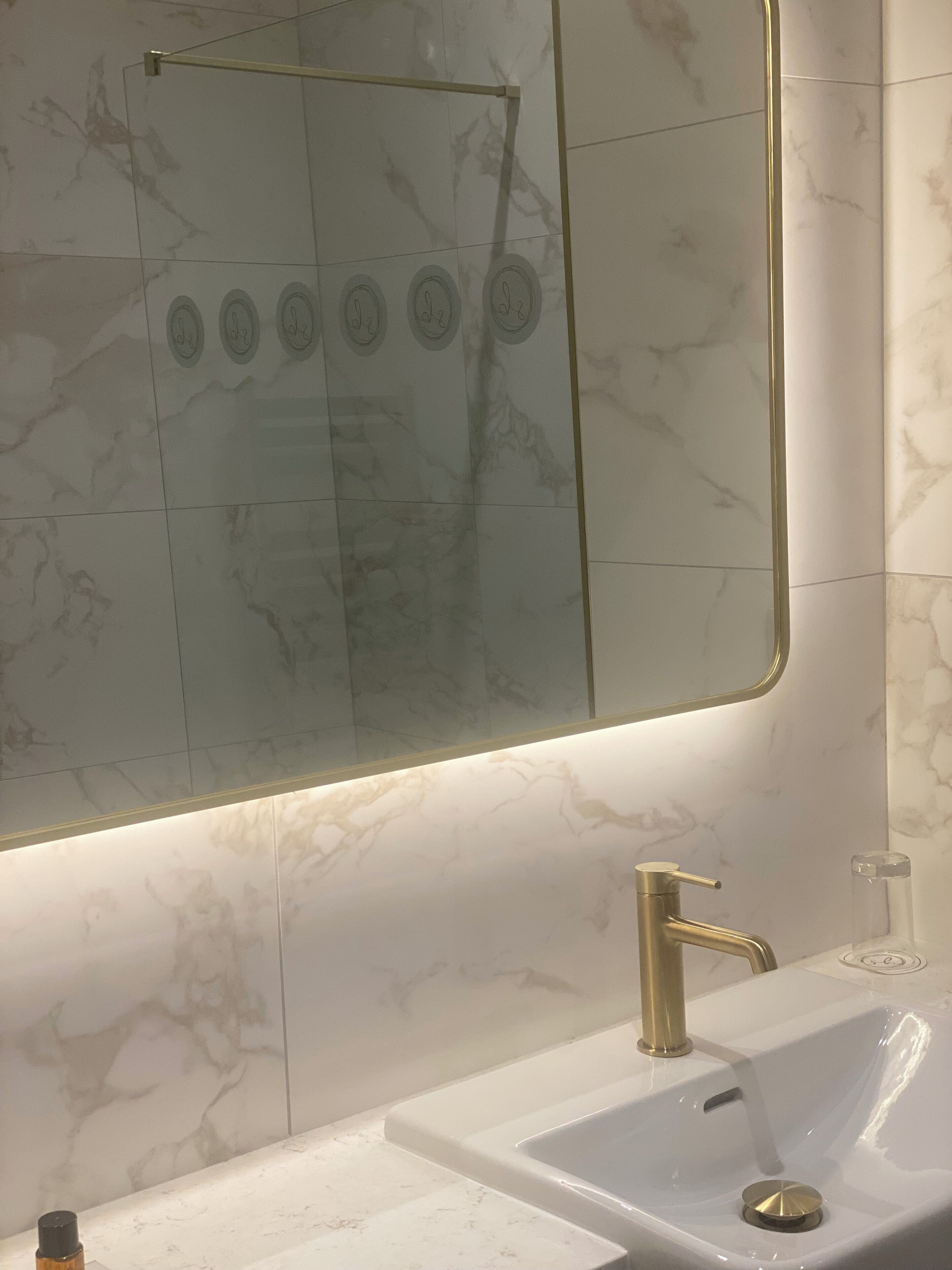 A bathroom with a marble-tiled wall, a large mirror, a gold faucet, and a white sink.