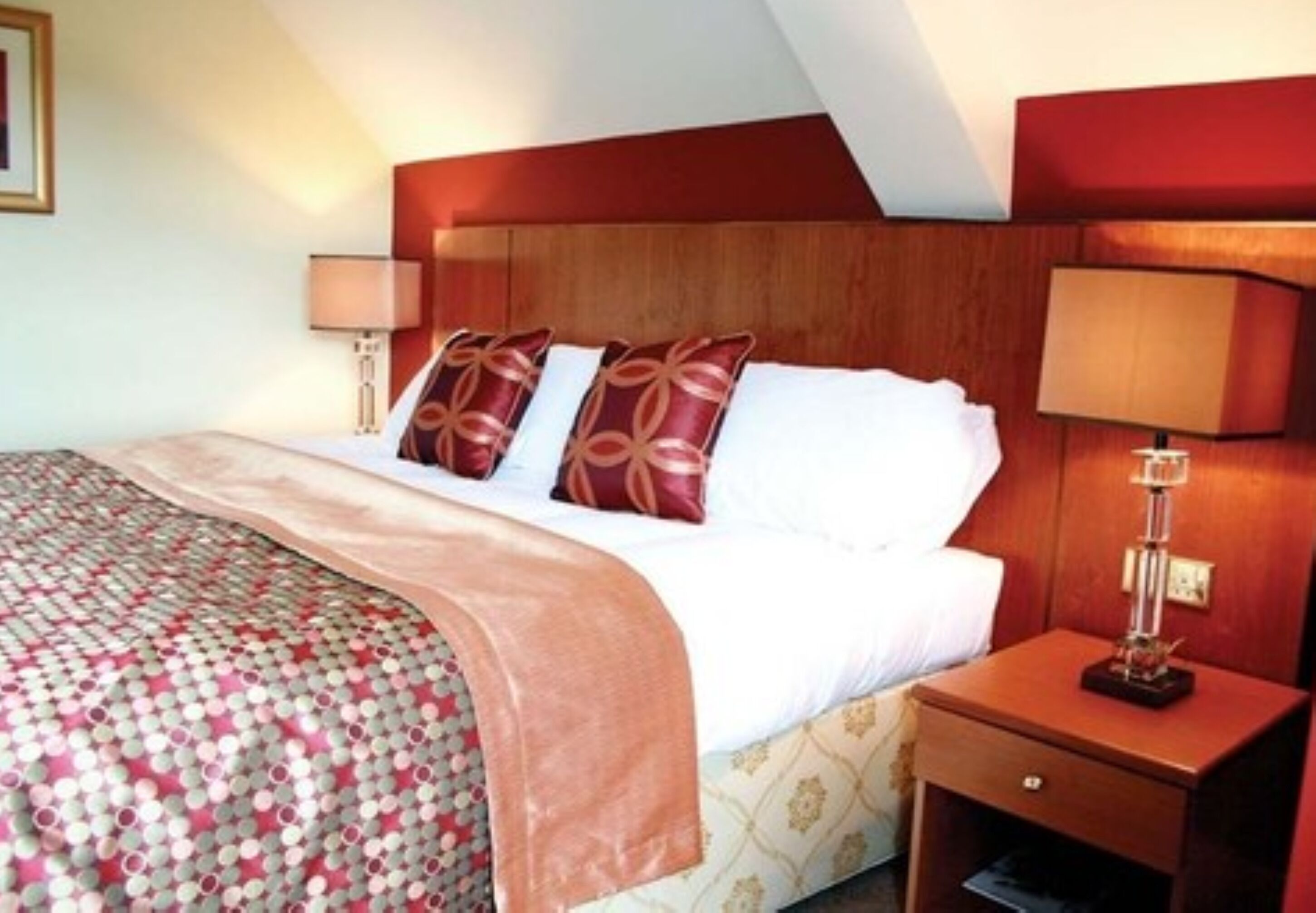 A hotel room with a wooden headboard, a bed with white linens and two decorative pillows, a bedside table with a lamp, and a patterned bedspread.