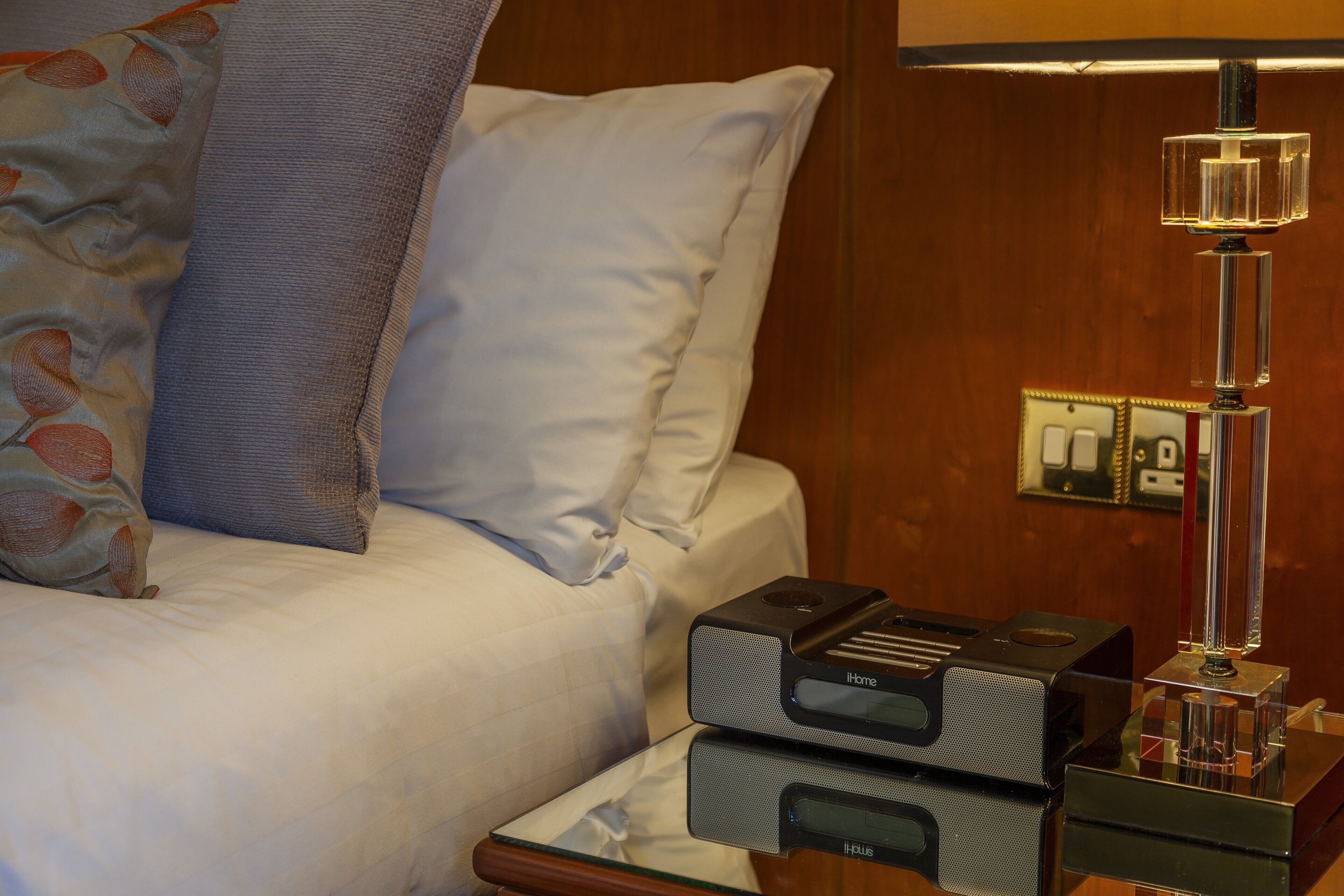Una cama con sábanas blancas y almohada azul, una mesita de noche con lámpara, y dos dispositivos electrónicos con la etiqueta "iBus" sobre una pared de madera.