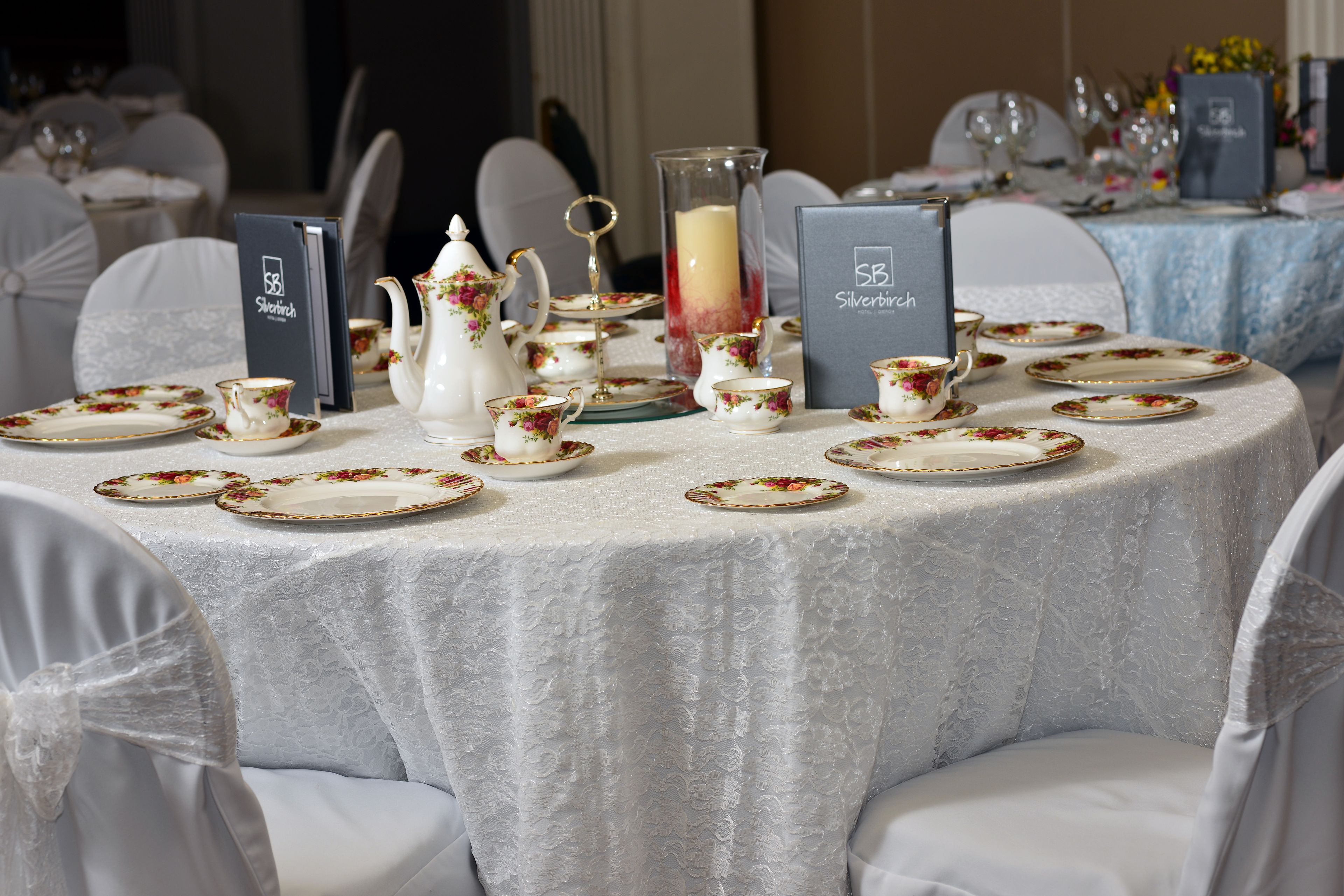 Une table ronde dressée pour un événement formel, avec de la vaisselle ornée de motifs floraux, un centre de table composé de bougies et un présentoir à menu.