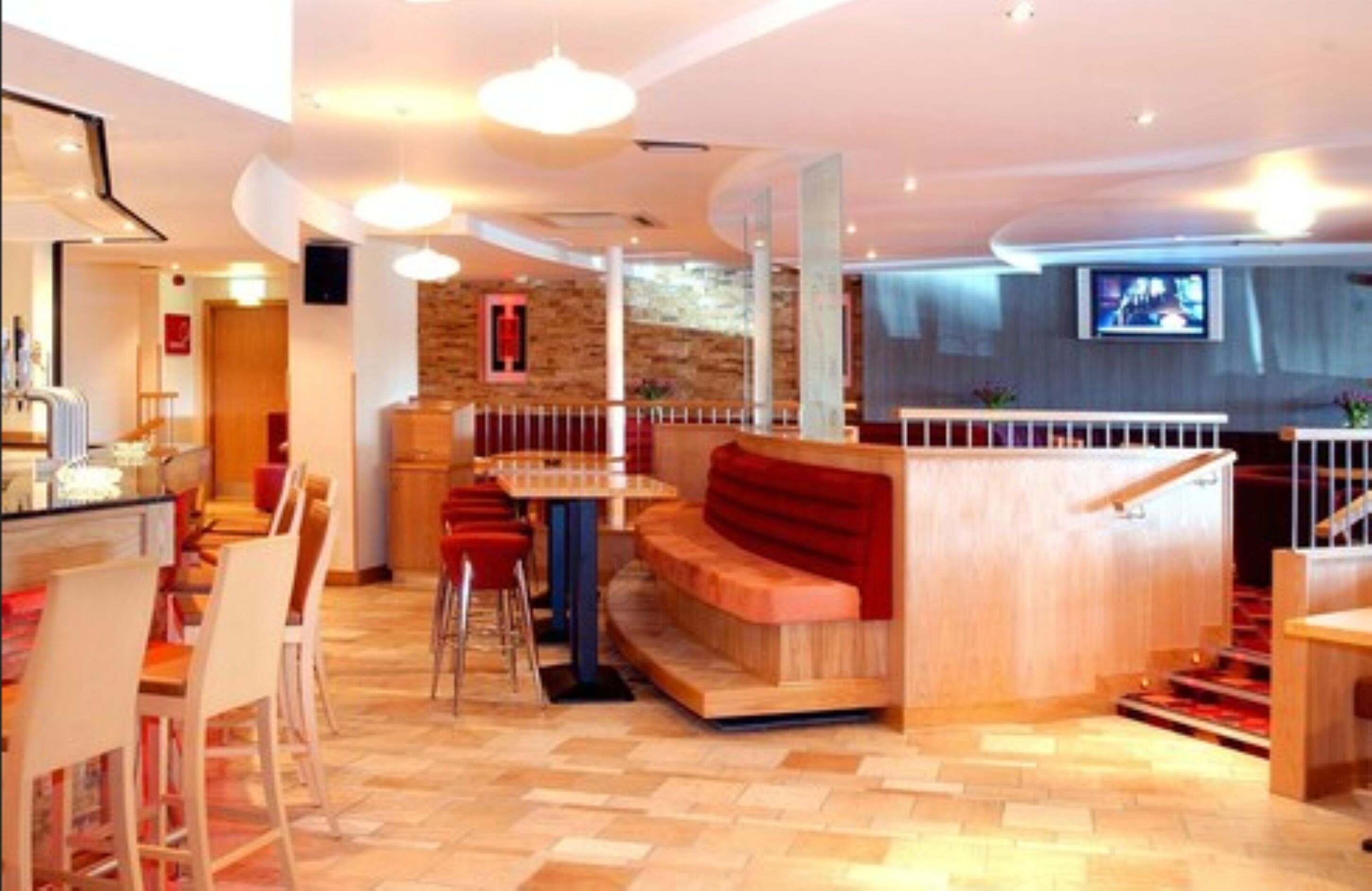 Un hall d’hôtel moderne avec un comptoir d’accueil en bois, un espace salon avec des bancs rouges et un escalier recouvert d’une moquette rouge.