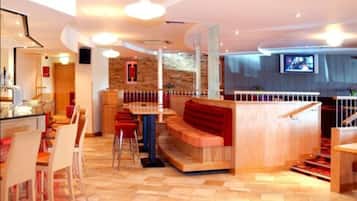 Un hall d’hôtel moderne avec un comptoir d’accueil en bois, un espace salon avec des bancs rouges et un escalier recouvert d’une moquette rouge.