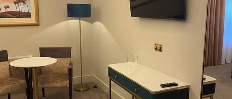 A hotel room with a round table, two armchairs, a desk with a mirror, a TV mounted on the wall, and a floor lamp.