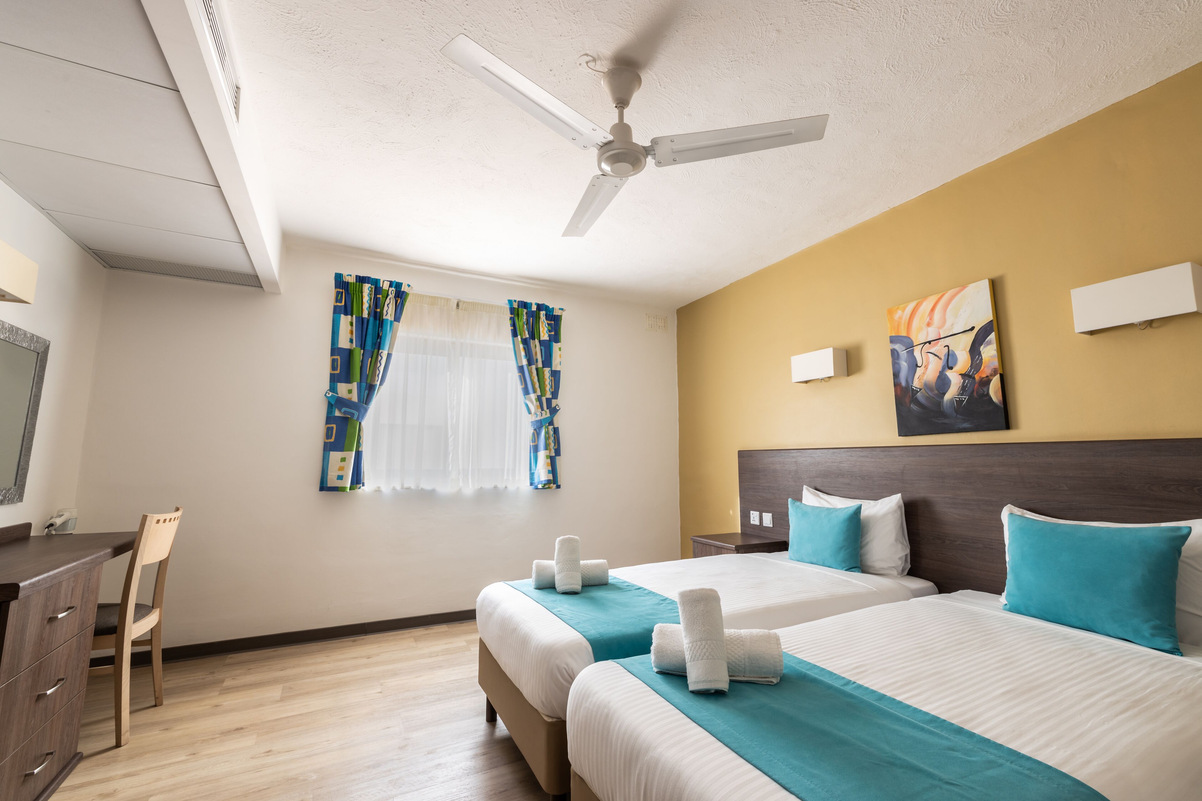 A hotel room with two beds, a desk, a chair, a ceiling fan, and artwork on the walls.