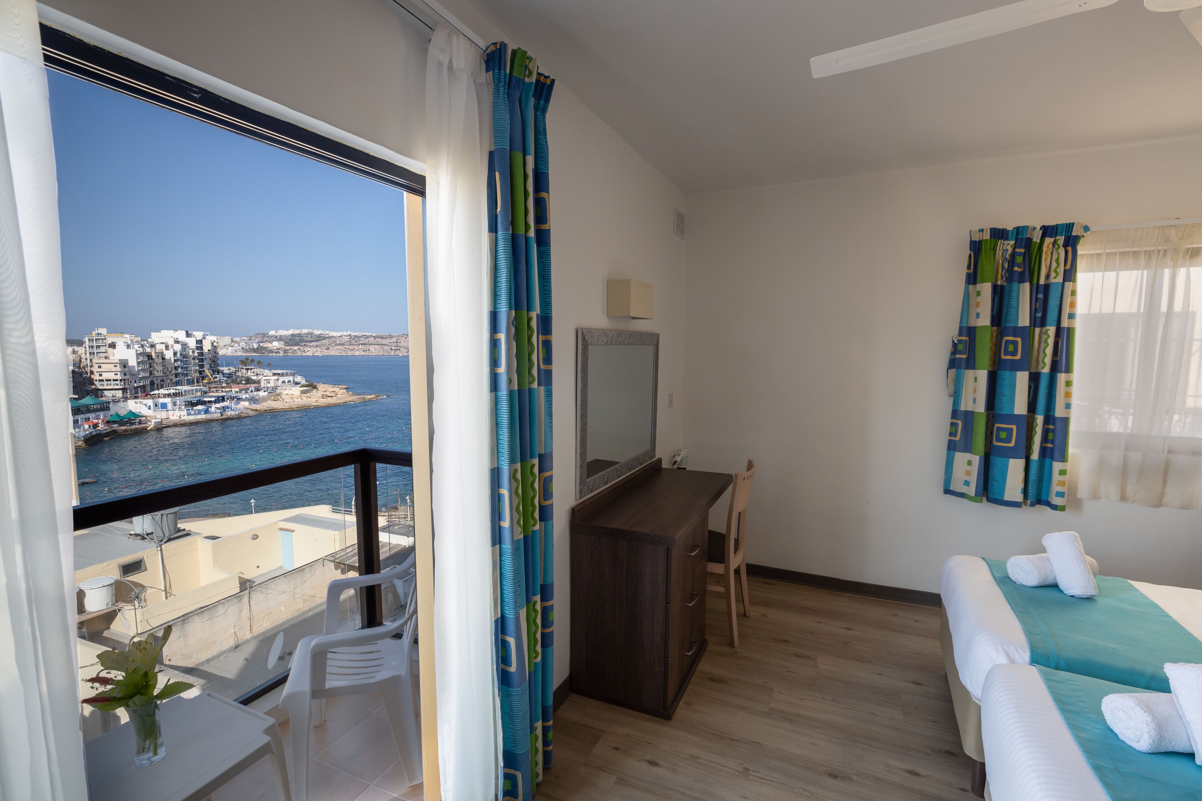 Habitación de hotel con balcón, escritorio, silla, cama con toallas y vista al mar y edificios.