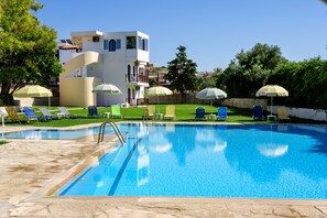A swimming pool with lounge chairs and umbrellas, surrounded by a white building with a balcony. - Prince Of Lillies (Hersonissos)